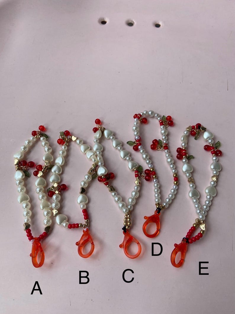 Peut inclure: Cinq lani&egrave;res perl&eacute;es avec des perles blanches, des perles en forme de c&oelig;ur, des accents rouges et des d&eacute;tails dor&eacute;s. Chaque lani&egrave;re a un fermoir orange vif. Les lani&egrave;res sont &eacute;tiquet&eacute;es A, B, C, D et E.