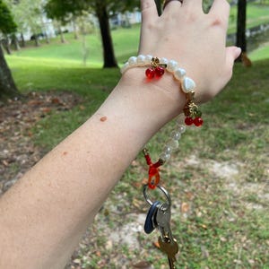 Peut inclure: Un bracelet de perles blanches avec des d&eacute;tails dor&eacute;s et des breloques de cerises rouges. Le bracelet est attach&eacute; &agrave; un porte-cl&eacute;s avec plusieurs cl&eacute;s. L'arri&egrave;re-plan est une sc&egrave;ne ext&eacute;rieure floue avec des arbres et de l'herbe.