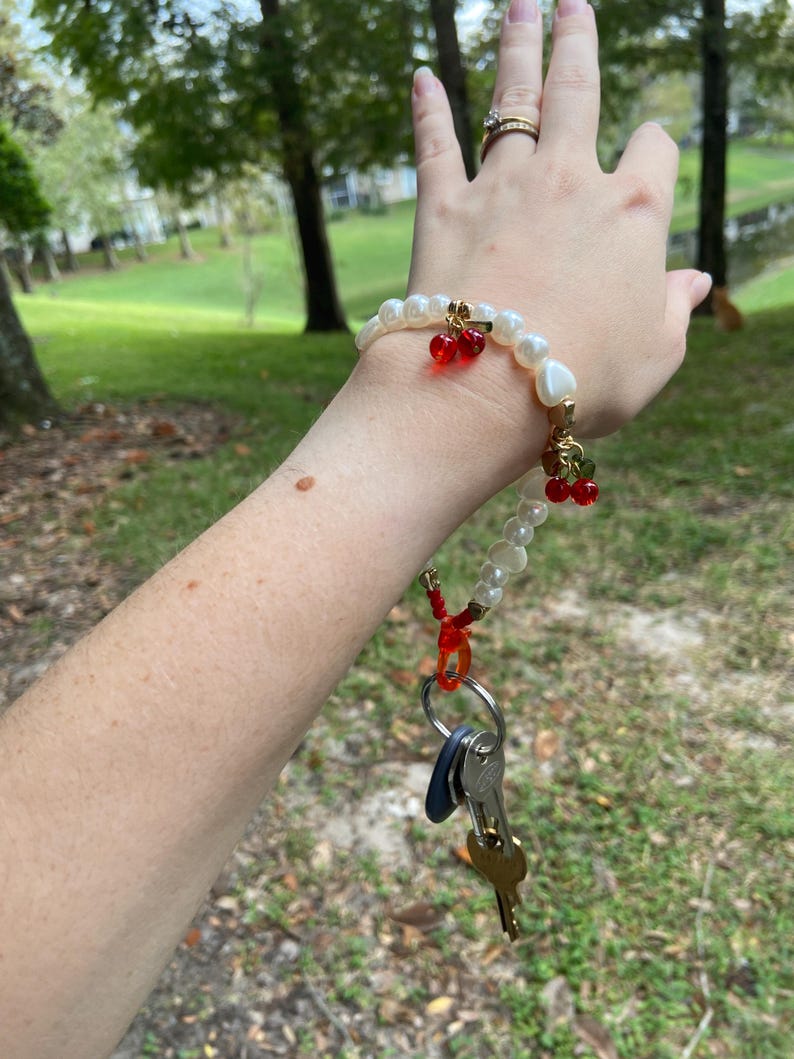 Peut inclure: Un bracelet de perles blanches avec des d&eacute;tails dor&eacute;s et des breloques de cerises rouges. Le bracelet est attach&eacute; &agrave; un porte-cl&eacute;s avec plusieurs cl&eacute;s. L'arri&egrave;re-plan est une sc&egrave;ne ext&eacute;rieure floue avec des arbres et de l'herbe.