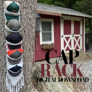 May include: A handmade gray macrame cap rack with several baseball caps hanging from it. The caps are various colors, including green, black, and red. The text "CAP RACK DIGITAL DOWNLOAD" is displayed on the image.