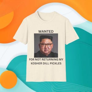 May include: A cream-colored short-sleeve t-shirt featuring a graphic print of a man wearing glasses. Above the image is the text WANTED, and below the image is the text FOR NOT RETURNING MY KOSHER DILL PICKLES in black capital letters.