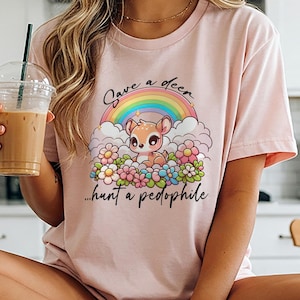 May include: A light pink t-shirt featuring a cartoon deer design with a rainbow and floral elements. The text on the shirt reads "Save a deer... hunt a pedophile."