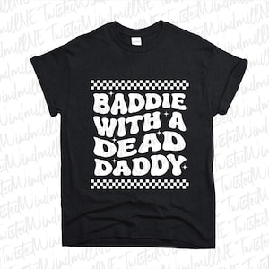 May include: Black t-shirt featuring the white text "BADDIE WITH A DEAD DADDY" in a retro font, with a checkered border. A casual, graphic tee.