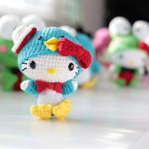 May include: A crocheted Hello Kitty toy dressed as a penguin. The toy is wearing a blue and white hat with a red bow, a red bow tie, and yellow feet.