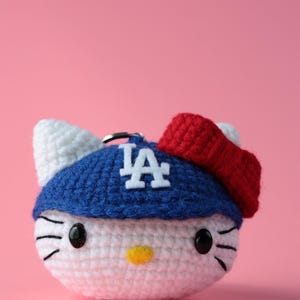 May include: A crocheted keychain featuring a white, round character with black eyes and whiskers, a yellow nose, and white ears. It wears a blue hat with the white letters "LA" and a red bow. The background is a solid pink.
