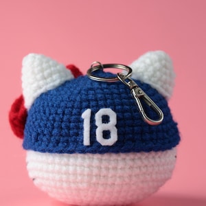 May include: A handmade crochet keychain in the shape of a cat's head. The keychain features a blue and white color scheme with white ears, a red accent, and the number 18 in white. It includes a silver keyring.