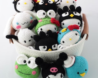 May include: A collection of handmade crochet character keychains in a white basket. The keychains feature various characters in white, black, green, and blue, with red bows and accents. The characters include a frog, penguin, and other popular designs.
