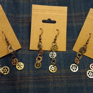 Peut inclure: Trois paires de boucles d'oreilles steampunk dorées, argentées et bronze avec des breloques en forme d'engrenages suspendus à un crochet. Les boucles d'oreilles sont présentées sur du carton brun.