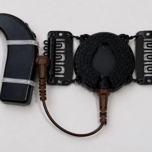 Pode incluir: Um aparelho auditivo retroauricular preto ligado por um fio castanho a um dispositivo circular preto com dois ganchos de cabelo pretos de cada lado. O aparelho auditivo tem um gancho transparente e está preso com duas fitas de plástico.