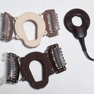 Könnte beinhalten: Drei Haaraccessoires: zwei mit Clips und einem U-förmigen Mittelteil sowie ein kreisförmiges Teil mit einem Kabel. Die Accessoires sind in Braun- und Beigetönen gehalten und haben silberfarbene Metallclips.