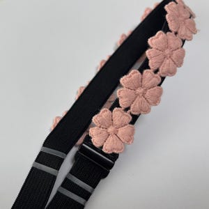 Peut inclure: Sangle élastique noire avec des fleurs roses brodées. La sangle est ornée d'une boucle en plastique noir et de bandes argentées réfléchissantes. Les fleurs sont espacées le long du bord.