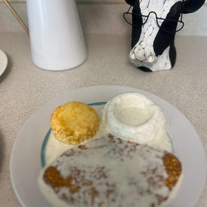 May include: A plate of biscuits and gravy with a golden biscuit, a large piece of fried chicken, and a small bowl of gravy. A black and white cow head figurine with glasses is in the background.