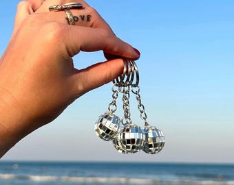 Può includere: Un set di tre portachiavi a sfera da discoteca. Ogni portachiavi presenta una piccola sfera da discoteca sferica con piastrelle a specchio riflettenti, attaccata a una catena e un portachiavi argentati. Lo sfondo mostra una spiaggia e un cielo blu.