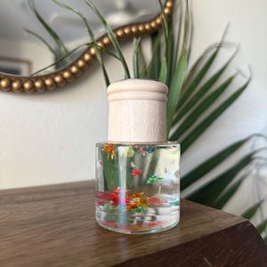 Könnte beinhalten: Kleine, klare Glasflasche mit hellem Holzdeckel. Die Flasche ist mit einer klaren Flüssigkeit und bunten, winzigen Blumen gefüllt. Die Flasche steht auf einer Holzoberfläche, mit einem Spiegel und Grünzeug im Hintergrund.