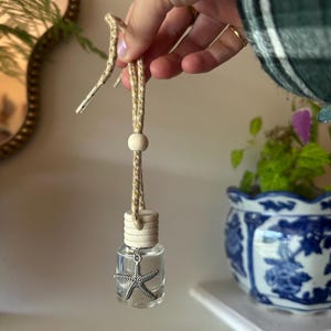 Puede incluir: Una pequeña botella de vidrio transparente con tapa de madera clara, suspendida por un cordón beige moteado con una cuenta de madera. Un dije de metal plateado en forma de estrella de mar está sujeto al cuello de la botella con líquido.