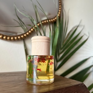 May include: A small, clear glass bottle filled with yellow liquid and small red and green floral elements. The bottle has a light wood cap. The background includes a gold-framed mirror and green foliage.