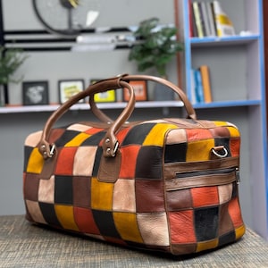 Op de afbeelding: Een patchwork leren reistas met bruine, zwarte, rode, gele en beige vierkanten. De tas heeft twee bovenste handvatten en een ritsvak aan de zijkant.