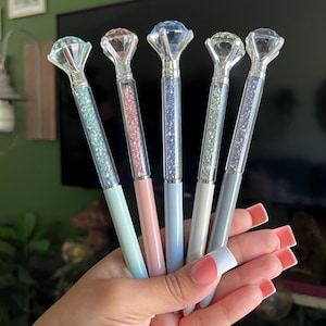 May include: Five pens with clear barrels filled with small, colorful crystals and topped with large, faceted diamond-shaped toppers. The pens feature pastel-colored lower bodies in light green, pink, light blue, cream, and gray, with silver accents.