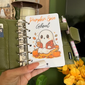 May include: A green leather planner with a decorative insert featuring a cartoon ghost holding a pumpkin spice latte. The insert reads "Pumpkin Spice Ghoul" with fall-themed illustrations of pumpkins and leaves.