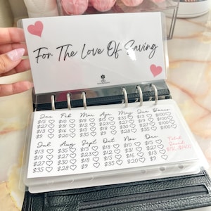 May include: A white planner with a clear cover and black rings. The planner has a page with a pink heart and the text "For The Love Of Saving". The page is a monthly budget tracker with the months of the year listed across the top and dollar amounts listed below each month.