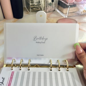 May include: A white plastic page protector with the text "Birthdays Sinking Fund" and "Daily Budgets" printed on it. The page protector is designed to fit a standard size planner.