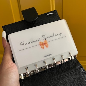 May include: Black leather planner with a white insert labeled "Personal Spending" in script font. A peach bow graphic is centered below the text. The planner is open, revealing silver binder rings.