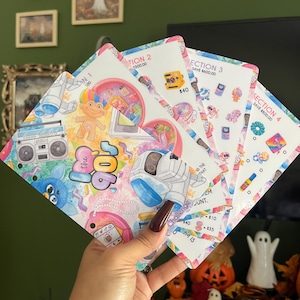 May include: A set of four white savings challenge cards featuring colorful 90s-themed illustrations including a boombox, a troll doll, a robot, and various retro icons. Each card is labeled with section numbers and dollar amounts for tracking savings.