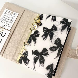 May include: A white planner with gold rings and a black and white bow pattern insert. The insert has the text "Dream" written on it. The planner has a quote from Jeremiah 29:11 on a small card.