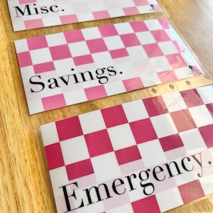 May include: Three rectangular clear plastic cash envelopes featuring a pink and white checkered pattern. Each envelope is labeled with black text reading Misc., Savings., and Emergency. The envelopes have punched holes along the side for binding.