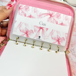May include: Pink zippered wallet with gold hardware, open to reveal a clear card holder with a pink bow pattern and the word "Gas". The wallet also has a blank white notepad with gold rings.