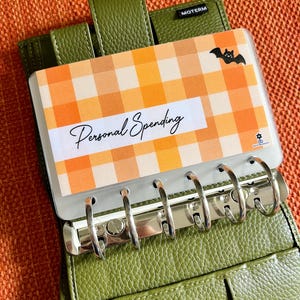 May include: An olive green textured leather binder with a six-ring metal mechanism. A clear plastic divider is inserted, featuring an orange and yellow gingham pattern, a small black bat graphic, and the text Personal Spending in black script.