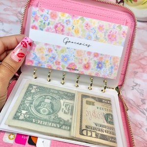May include: A pink zippered wallet with a floral patterned insert labeled "Groceries." The wallet is open, revealing cash and clear plastic dividers. The wallet is designed for budgeting and organizing finances.