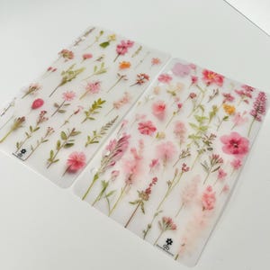 May include: Two clear plastic sheets with pressed pink and white flowers. The sheets are designed for use in a planner or journal.