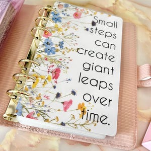 May include: A white planner with a floral design and the quote "Small steps can create giant leaps over time." The planner has gold rings and is closed.