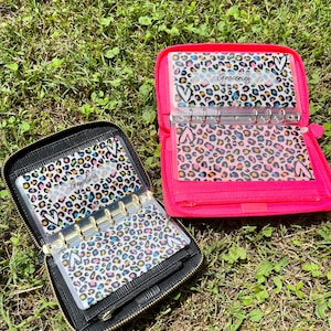 May include: Two zippered budget binders on grass. One is black, the other is bright pink. Both have clear inserts with a leopard print design in multiple colors. The black binder has "Gasoline" written on one of the inserts. The pink binder has "Groceries" written on one of the inserts.