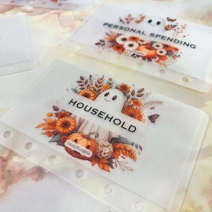 May include: A set of four clear plastic budget tracker inserts with a fall theme. Each insert features a ghost, fall leaves, pumpkins, and sunflowers. The text on the inserts reads "HOUSEHOLD", "PERSONAL SPENDING", and "DailyBudgets".