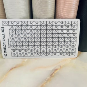 May include: A white 100 envelope challenge tracker with black grid lines and numbers 1 through 100. The text "100 ENVELOPE CHALLENGE" and "DailyBudgets" is printed on the left side of the tracker.
