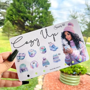May include: A white rectangular card featuring the text Cozy Up and Saves $60. The card displays illustrations of winter items including slippers, a mug, a candle, boots, a sweater, earmuffs, a beanie, and a stack of folded sweaters with prices.