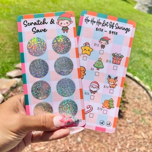 May include: Two colorful scratch-off cards are held in a hand. One card reads "Scratch & Save" with holographic circles. The other card says "Ho Ho Ho Lot Of Savings" with Christmas-themed illustrations and amounts.