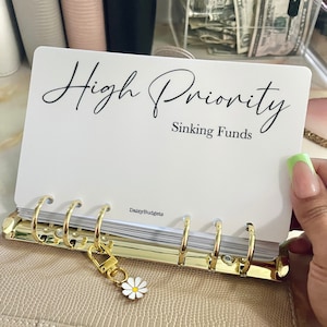May include: White card with black text that reads "High Priority Sinking Funds" with the text "DaisyBudgets" in the bottom right corner. The card is in a gold binder with a daisy charm.