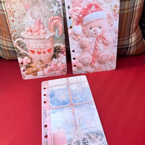 May include: Three decorative planner inserts with winter and holiday themes. One features a pink mug of hot chocolate with marshmallows and a candy cane. Another shows a pink teddy bear in a Santa hat holding a candy cane. The third depicts a snowy window and a lit candle.