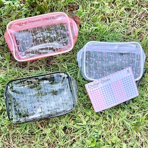 May include: Three rectangular clear pouches with zipper closures, featuring a repeating bow pattern. One pouch has pink trim, one has light blue trim, and one has black trim. A rectangular card labeled Dollar Dots is placed near the blue pouch.