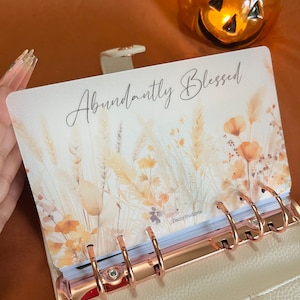 May include: A cream-colored planner with a gold ring binder. The cover features a watercolor design of fall foliage and the text "Abundantly Blessed". The planner is open to a page with the text "DaisyBudgets".