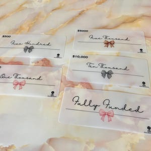 May include: Four gift cards with different amounts: $500, $1000, $5000, and $10,000. Each card has a pink ribbon and the amount written in black text. The last card says "Fully Funded" in black text.