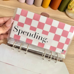 May include: A rectangular plastic cash envelope for a binder featuring a pink and white checkered pattern. The word Spending. is printed in black serif font on the front. A small black flower logo with the letters db is in the bottom corner.