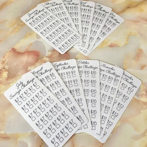 May include: A set of 12 white printable savings challenge cards for each month of the year. Each card has a black border and a black check box for each day of the month. The cards are labeled with the month and the words "Savings Challenge".