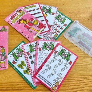 May include: A collection of pink and green Christmas-themed cards and a clear plastic pouch. The cards feature the Grinch, candy canes, and gift illustrations. The text on the cards includes "Merry Grinchmas" and savings challenges.