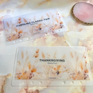 May include: Two clear plastic pages with a floral design and the text "CAROLINA CLASSIC FAIR Sinking Fund" and "THANKSGIVING Sinking Fund". The text "DaisyBudgets" appears at the bottom of each page.