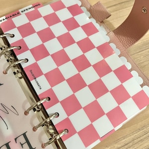 May include: A pink and white checkered planner divider with a scalloped edge, featuring the text DAISYBUDGETS printed on the side. The divider is secured within a six-ring metal binder mechanism inside a textured light pink leather cover.