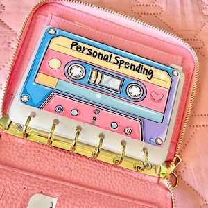 May include: A pink zippered binder featuring a six-ring gold metal mechanism. Inside, a white plastic insert displays a colorful illustration of a retro cassette tape with the text Personal Spending printed in black across the top center.